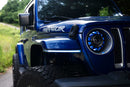 Blue Jeep Wrangler with 'Wrangler' branding and Morimoto fender chop kits with XRGB lighting on a road with greenery in the background