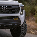 Close-up of a 2024 Toyota Tacoma truck's front grille equipped with Baja Designs lighting upgrades and bumper with a blurred natural background