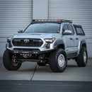 Toyota Tacoma with lifted suspension and an RTS Front Winch Bumper in front of a gray building.