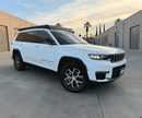 White Jeep Grand Cherokee with a prinsu pro roof rack parked on a concrete surface with buildings and palm trees in the background.